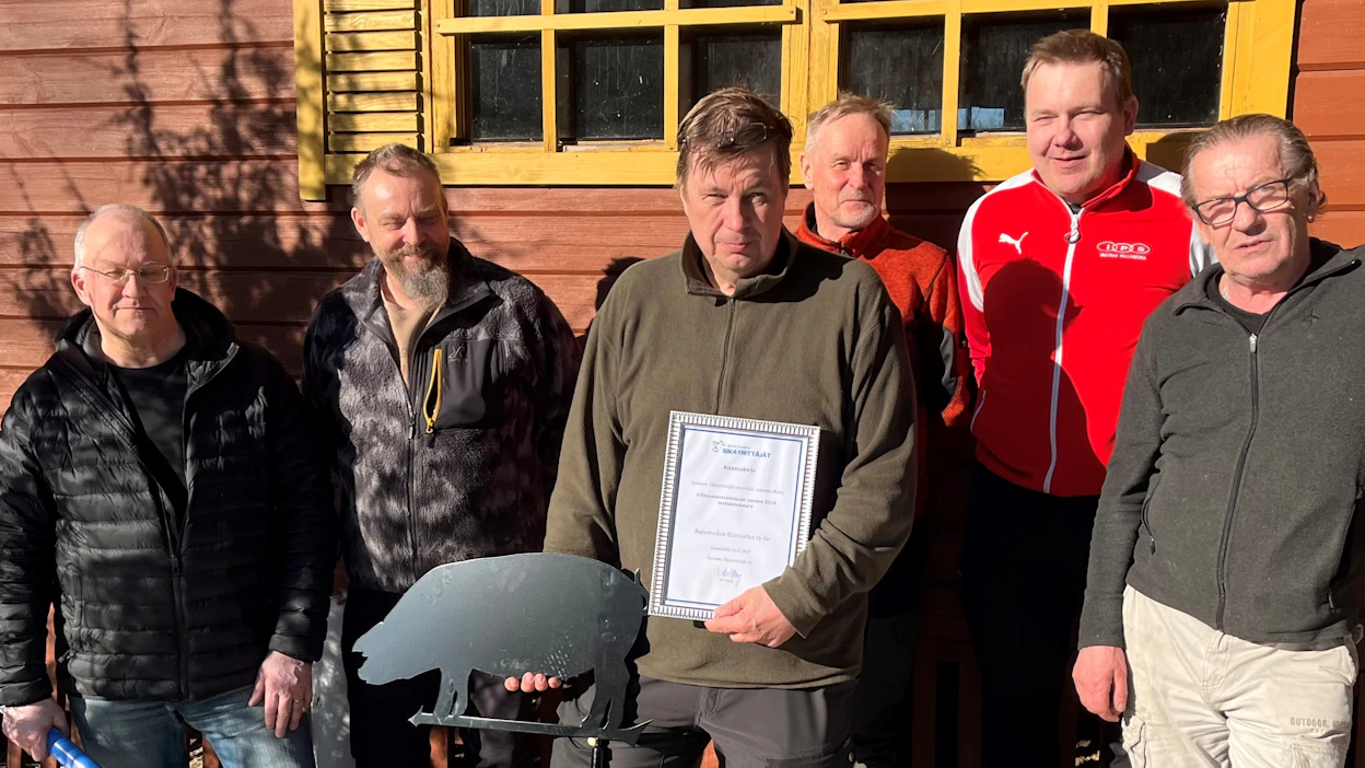 Villisianmetsästäjät kirkkaassa auringonpaisteessa metsästysmajan seinustalla. Vasemmalta Jouni Lauren, Terho Maksimainen, Mika Haaranen (pj.), Olli Ahonen, Ari Kankkunen ja Ari Sjöholm.