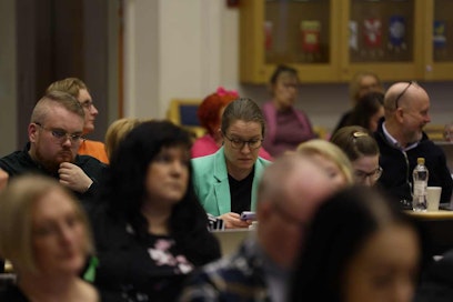 Vihreätakkinen nainen selaa puhelinta aluevaltuuston kokouksessa muiden kokoustajien keskellä