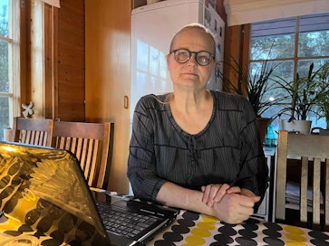 Henkilö istuu keittiössä pöydän ääressä kannettavan tietokoneen kanssa. Taustalla näkyy ikkunoista,  luonnonvaloa, sekä huonekasveja ja jääkaappi.