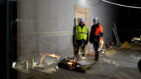 Kaksi miestä seisoo huoneessa ja taustalla rakennustyömaa.