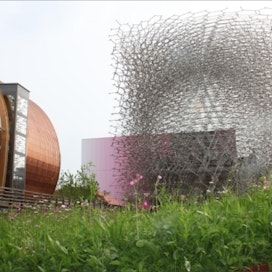 Britannian paviljongin alumiininen mehiläispesä on yhteydessä Britanniassa sijaitsevaan oikeaan mehiläispesään suristen sen kanssa yhteen ääneen.  Öisin keinopesä valaistaan led-lampulla. MErja Ojansivu