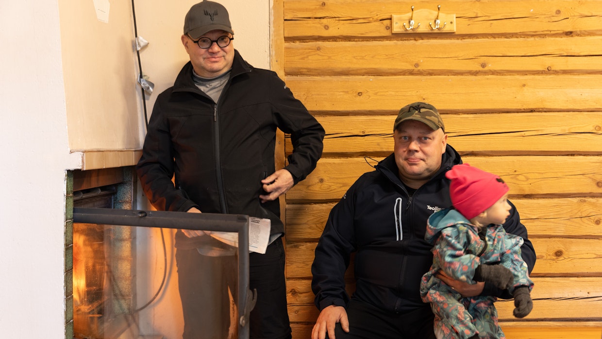 Kylätalo lämpiää puulla ja ilmalämpöpumpuilla. Risto Hopponen ja Taina (sylissä) seuraavat vieressä, kun Jarmo Tupasela laittoi tulet uuniin.