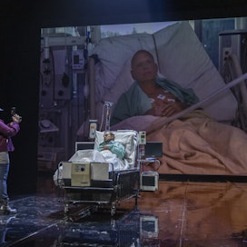 Mikko Pörhölä (Aleksandr Litvinenko) makaa sairaalavuoteessa. Taustalla kuva aidosta tilanteesta. Kameran takana Lumikki Väinämö.