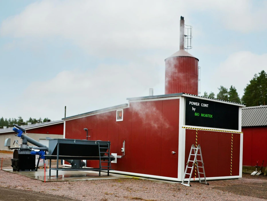 Siisti lämpökontti vanhan lämpölaitoksen vieressä on melko huomaamaton. Uuden laitoksen ominaisuudet parantavat kotieläintilan hygieniaa ja alentavat tuotantokustannuksia. Taustalla näkyy vanhan laitoksen rakenteita.