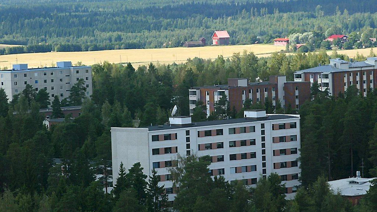 Kerrostaloja metsäisellä lähiöalueella. Taajaman takana pelto, jonka laidalla mm. kuivuri.