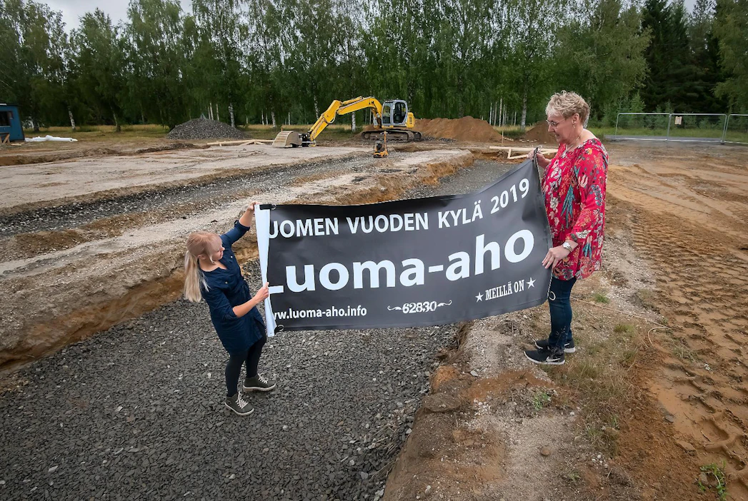 Luoma-ahon kyläyhdistyksen sihteeri Soili Mäki ja puheenjohtaja Sirkka Rajala esittelevät banderollia, jollainen tervehtii kylätiellä kulkijoita sunnuntaista lähtien. Kylätalon rakentaminen käynnistyi viime viikolla perustuskaivantojen teolla.