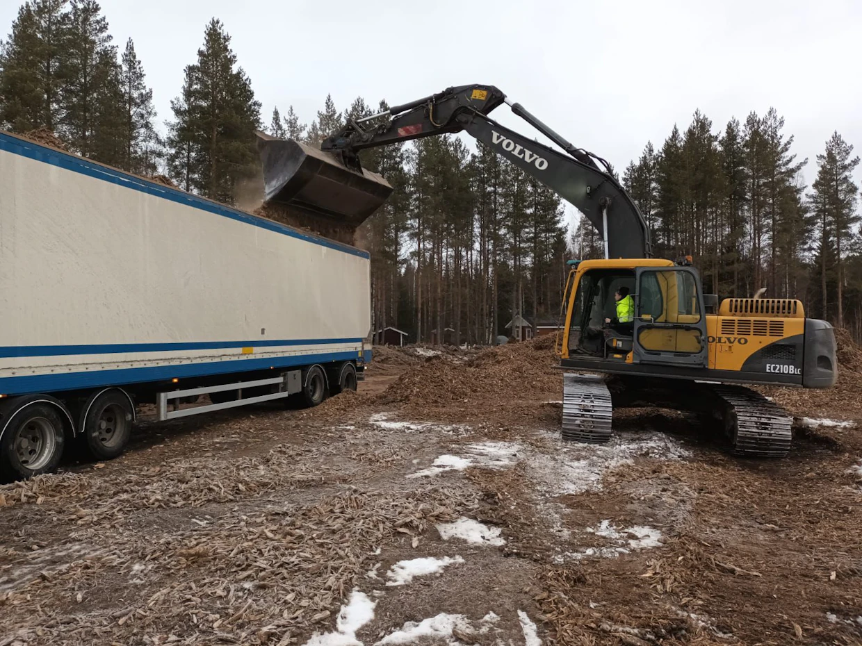 Iina Mäkinen kaivinkoneen puikoissa lastaamassa haketta yhdistelmäajoneuvoon.